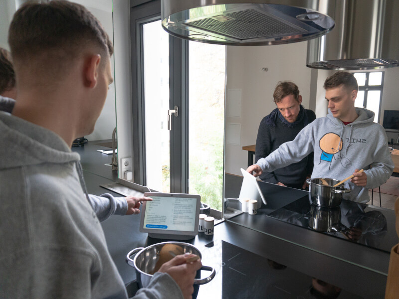 Zwei Personen bedienen den Google Nest Hub Max beim Kochen in der Küche mit dem Assistant