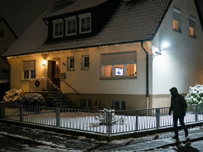 Einbrecher abschrecken: Zwielichtige Gestalt geht am Grundstück vorbei, sieht Überwachungskamera, eingeschaltetes Licht und Fernseher, halb heruntergelassene Rollläden und Flutlicht im Garten und geht weiter.