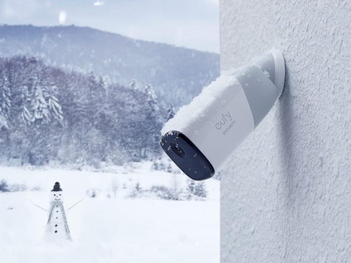 Bild einer weißen und dunkelgrauen Eufy-Überwachungskamera, die mit Schnee bedeckt ist und an einer weißen, strukturierten Wand befestigt ist, mit einer Winterlandschaft im Hintergrund.