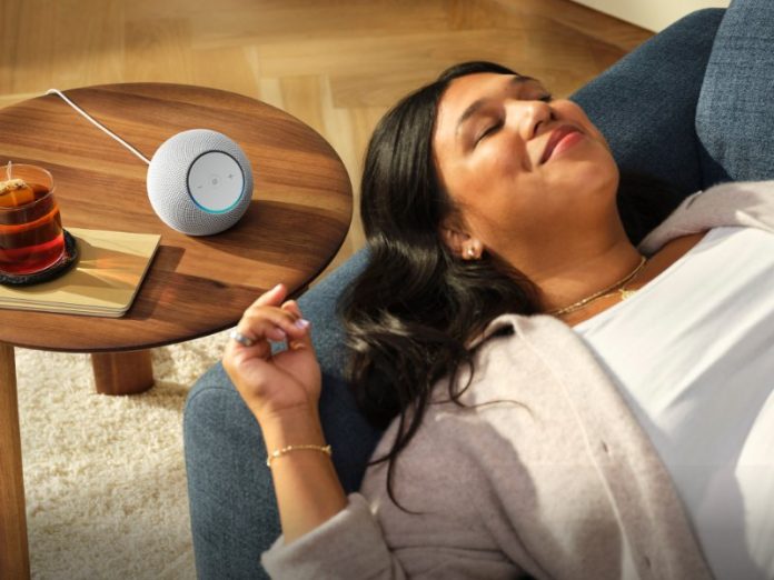 Eine Frau entspannt sich in einem blauen Sessel, während der weiße, kugelförmige Echo Dot mit blauem Lichtring auf einem Beistelltisch aus Holz neben einem Glas Tee steht.