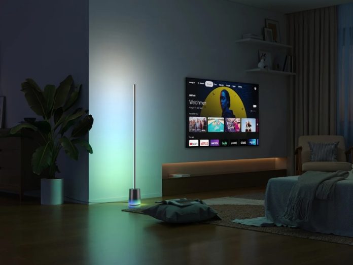 Gedimmtes Wohnzimmer mit intelligenter Govee-Stehleuchte, die einen blau-grünen Farbverlauf an die Wand neben einem Fernseher projiziert. Govee CES 2026 Stimmungsbild.
