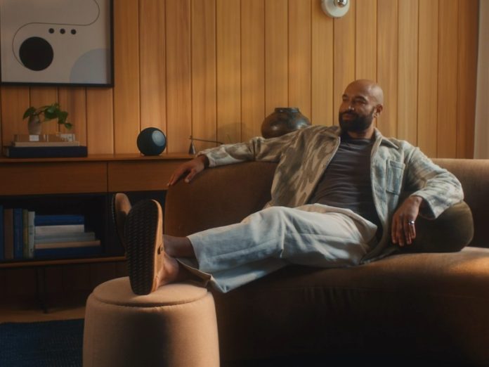 Ein Mann entspannt lässig auf einem braunen Sofa, während im Hintergrund ein dunkler, kugelförmiger Alexa Smart Speaker auf einem Holzregal steht. Dies veranschaulicht die Anwendung von Alexa für Einsteiger.
