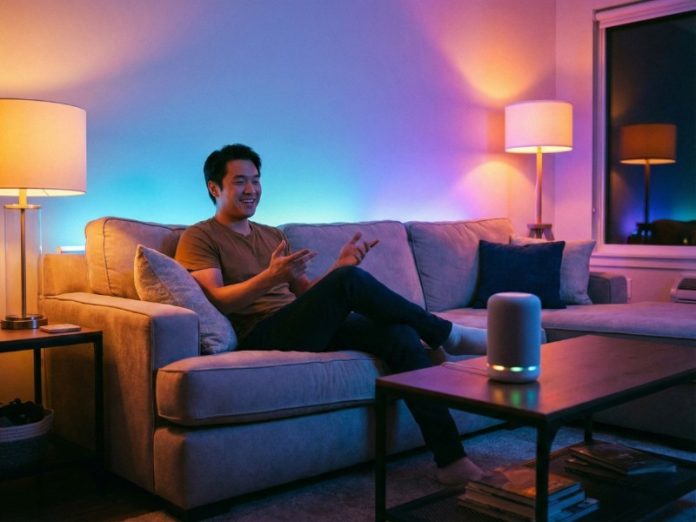 Mann auf der Couch mit smarten Lichtern im Wohnzimmer und Smart Speaker auf dem Couchtisch. Hier wird dargestellt, wie die Sprachsteuerung bei SmartThings funktioniert.