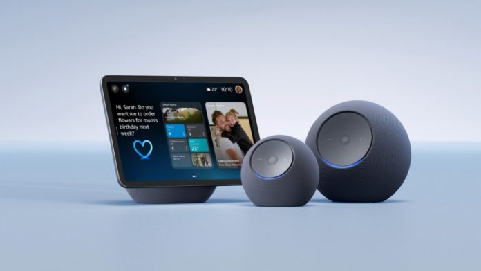 Drei dunkelgraue Amazon Alexa+ Deutschland Geräte – ein Echo Show Smart-Display mit personalisiertem Dashboard und zwei kugelförmige Echo-Lautsprecher mit charakteristischem blauem Lichtring – vor einem hellblauen Hintergrund.