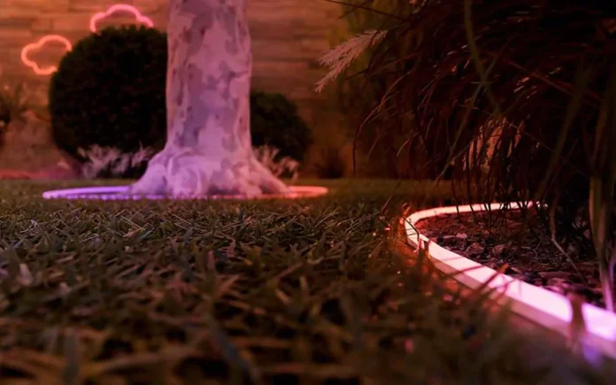 Philips Hue Neon Gradient Outdoor Lightstrip im Garten an der Rasenkante, Baum und Beleuchtung im Hintergrund.