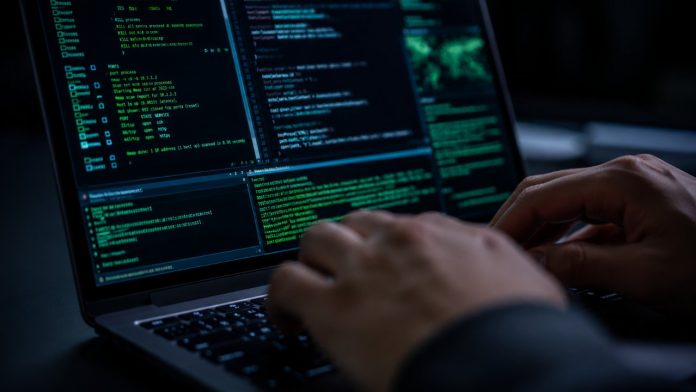 Jemand tippt auf einem Laptop mit mehreren Fenstern grünen und blauen Computercode auf den Bildschirm, was in einer dunklen Umgebung einen Datenleck in einem Smart-Home-Hotel symbolisiert.