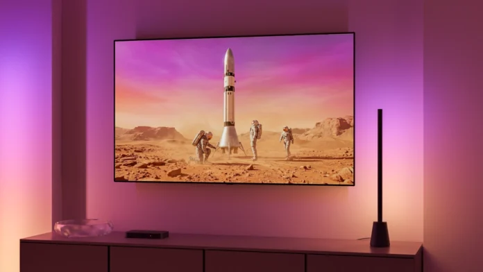 Philips Hue Play Gradient Tischleuchte neben dem Fernseher, auf dem ein bevorstehender Raketenstart gezeigt wird, die Tischleuchte leuchtet die Wand in zum Bild passenden Farben an.
