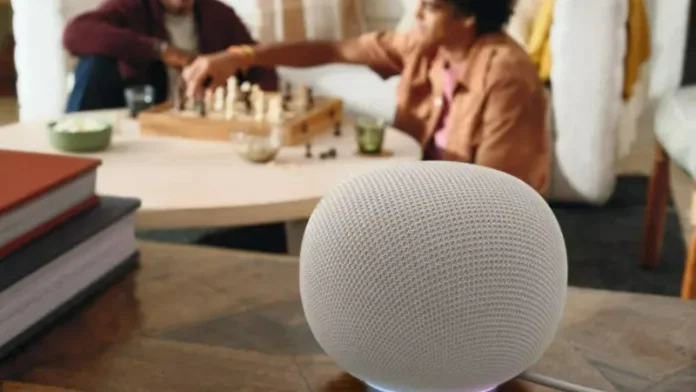 Der neue Google Home Speaker mit Gemini steht auf einem Holztisch im Vordergrund. Im verschwommenen Hintergrund spielen zwei Personen auf einer Bank Schach, dazwischen steht ein Couchtisch.