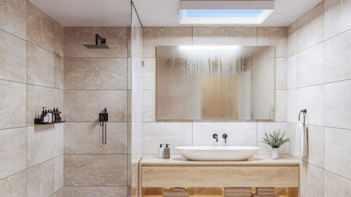 Ein modernes Badezimmer mit Fliesen in Beige in Marmoroptik, einem hellen Waschbeckenunterschrank aus Holz und einer Philips Skylight an der Decke, die ein hellblaues Licht verbreitet.