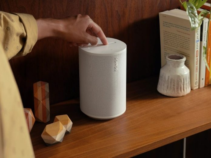 Sonos Verbindungsprobleme: Eine Hand drückt auf die Oberseite eines weißen Sonos-Lautsprechers auf einem dunkelbraunen Holzbrett, möglicherweise um ein Verbindungsproblem zu beheben. Rechts stehen Bücher und eine Vase, links dekorative Holzobjekte.