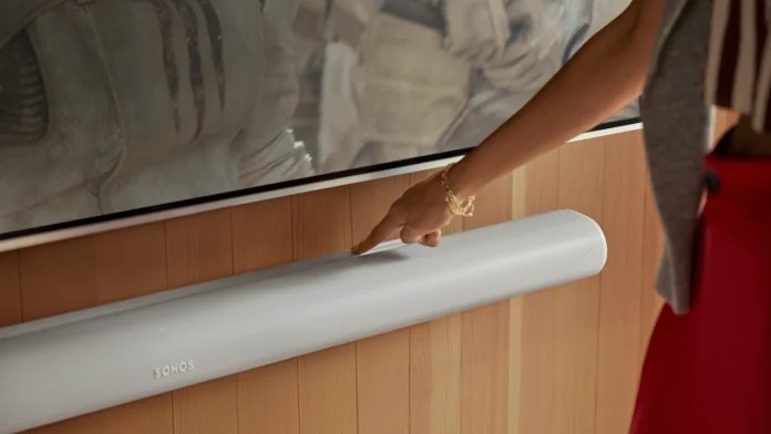 Eine Hand bedient die berührungsempfindlichen Tasten einer weißen Sonos-Soundbar, die an einer stilvollen Holzwand unter dem Fernseher angebracht ist.