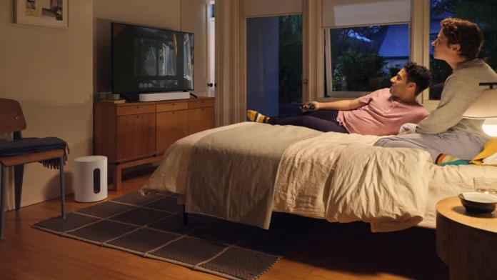 Ein Paar schaut entspannt fern in einem gemütlichen Schlafzimmer, in dem neben der hölzernen Kommode ein weißer Sonos Sub steht.