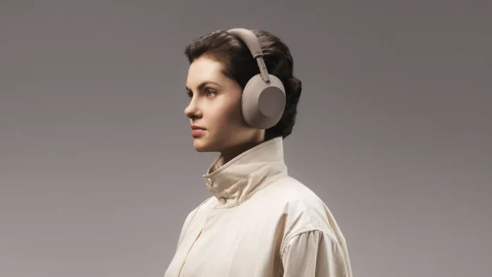 Eine Frau in einer hellen Jacke trägt die beigen Sony WH-CH730N-Kopfhörer mit Geräuschunterdrückung vor einem neutralen grauen Hintergrund.