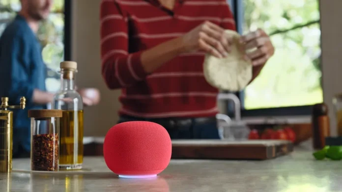 Ein roter Google Home-Lautsprecher mit Gemini AI steht auf einer Arbeitsplatte in der Küche, während im Hintergrund eine Person Teig zubereitet.