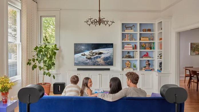 Familie auf blauer Couch im hellen Wohnzimmer schaut einen Film auf dem Fernseher. Umgeben sind sie von einem Sonos Heimkino-Set bestehend aus Arc Ultra, Sub und zwei Era 300.