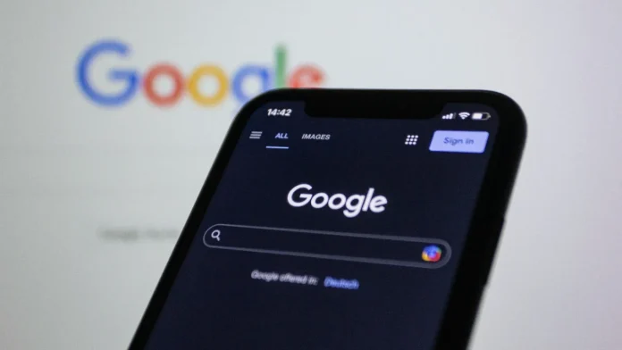 Smartphone mit der Google-Suchseite im Dunkelmodus vor einem unscharfen Google-Logo im Hintergrund.