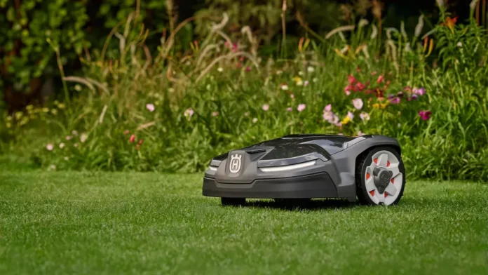 Ein grauer Husqvarna Automower 415X-Mähroboter mäht einen gepflegten grünen Rasen entlang einer üppigen Rabatte voller bunter Blumen.