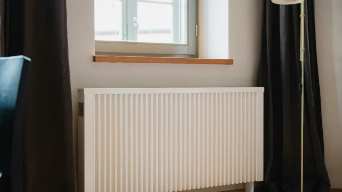 Ein weißer, moderner Heizkörper unter einer Fensterbank aus Holz in einem Zimmer mit dunklen Vorhängen.