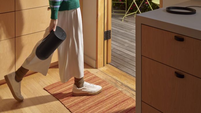Eine Person in einem grün-blau gestreiften Pullover und einer weiten weißen Hose trägt einen schwarzen Sonos Move-Lautsprecher aus einer hellen Holzküche auf eine sonnenüberflutete Terrasse mit hellgrünen Gartenmöbeln.