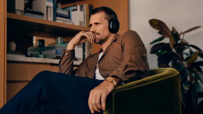 Nachdenklicher Mann mit schwarzem Sonos-Kopfhörer in einem grünen Sessel vor einem Bücherregal.
