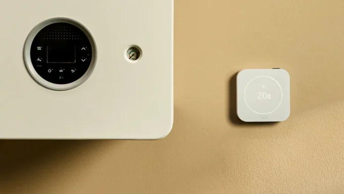 Ein tado-Smart-Thermostat neben einem weißen Heizkessel an einer beigen Wand.