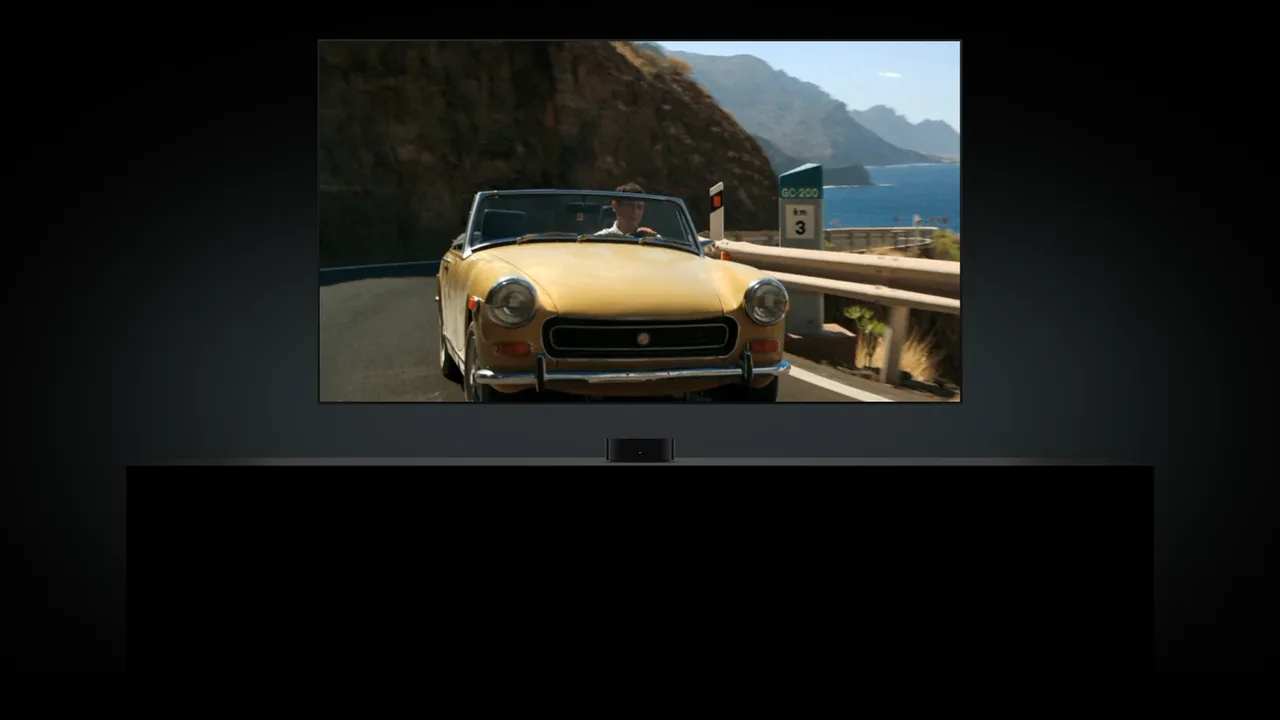 Apple TV 4K an einem Fernseher mit einer Filmszene: ein gelbes Cabrio auf einer Küstenstraße.