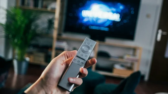 Hand hält eine Fernbedienung vor einem Fernseher im Wohnzimmer