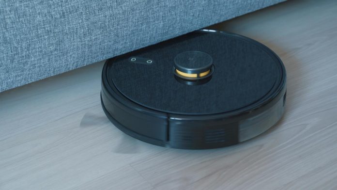 Schwarzer Saugroboter fährt unter einem Sofa über einen Holzboden