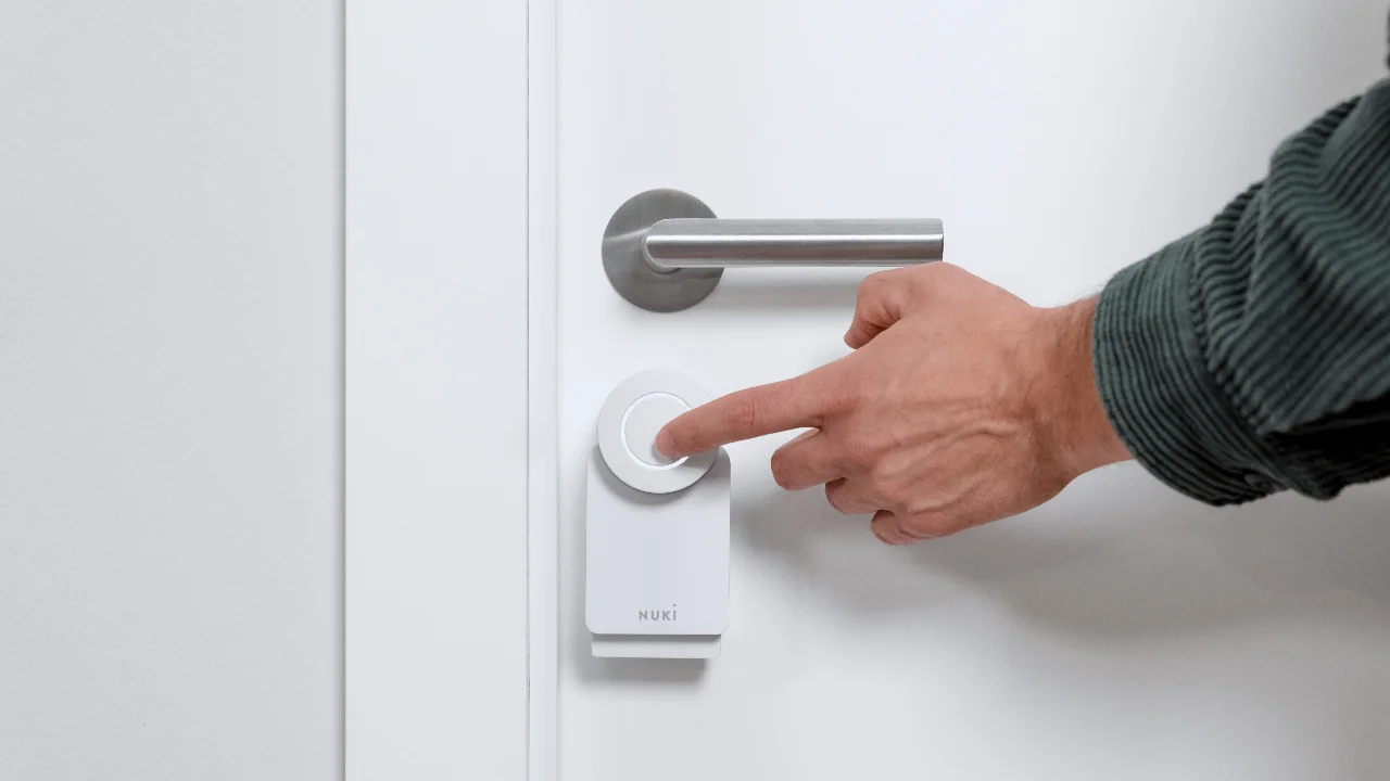Eine Hand drückt auf den Knopf eines weißen Nuki Smart Locks an einer weißen Tür.